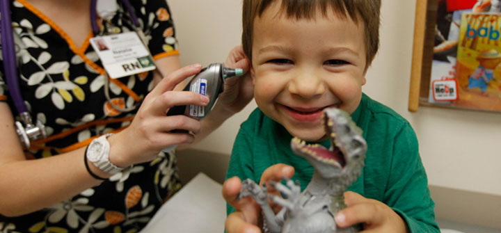 Smiling boy holding dinosaur while nurse checks his ears